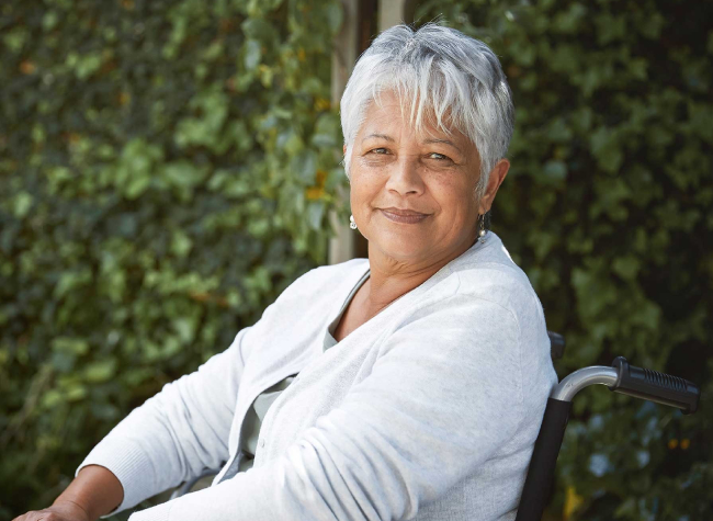 A headshot of a grey-haired woman of South Asian descent, sitting in a wheelchair and smiling softly at the camera.